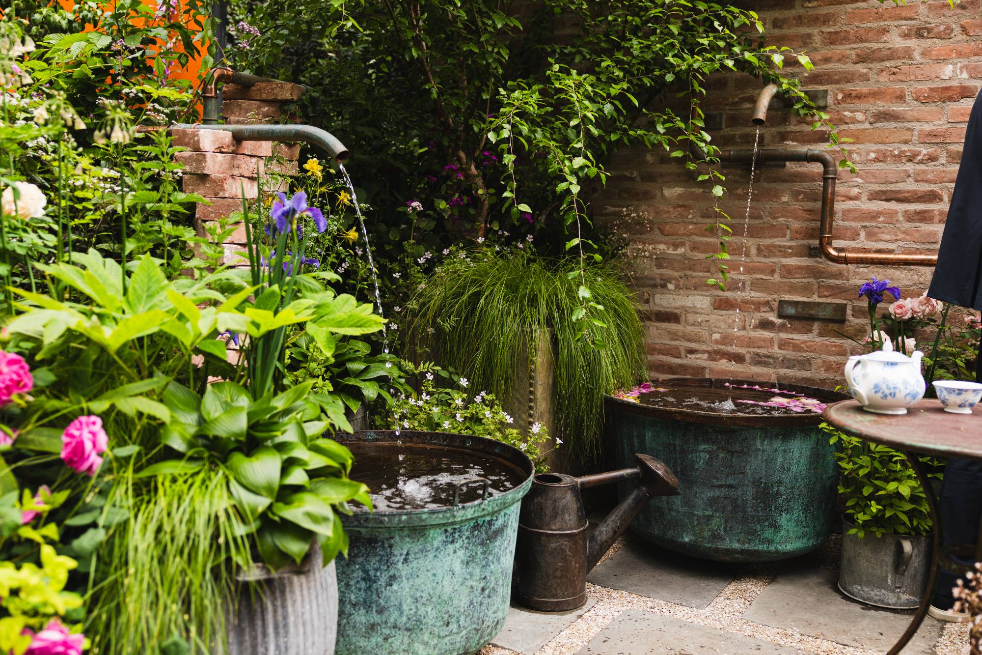 Garden scene with potted plants, a brick wall, and a table with a teapot.